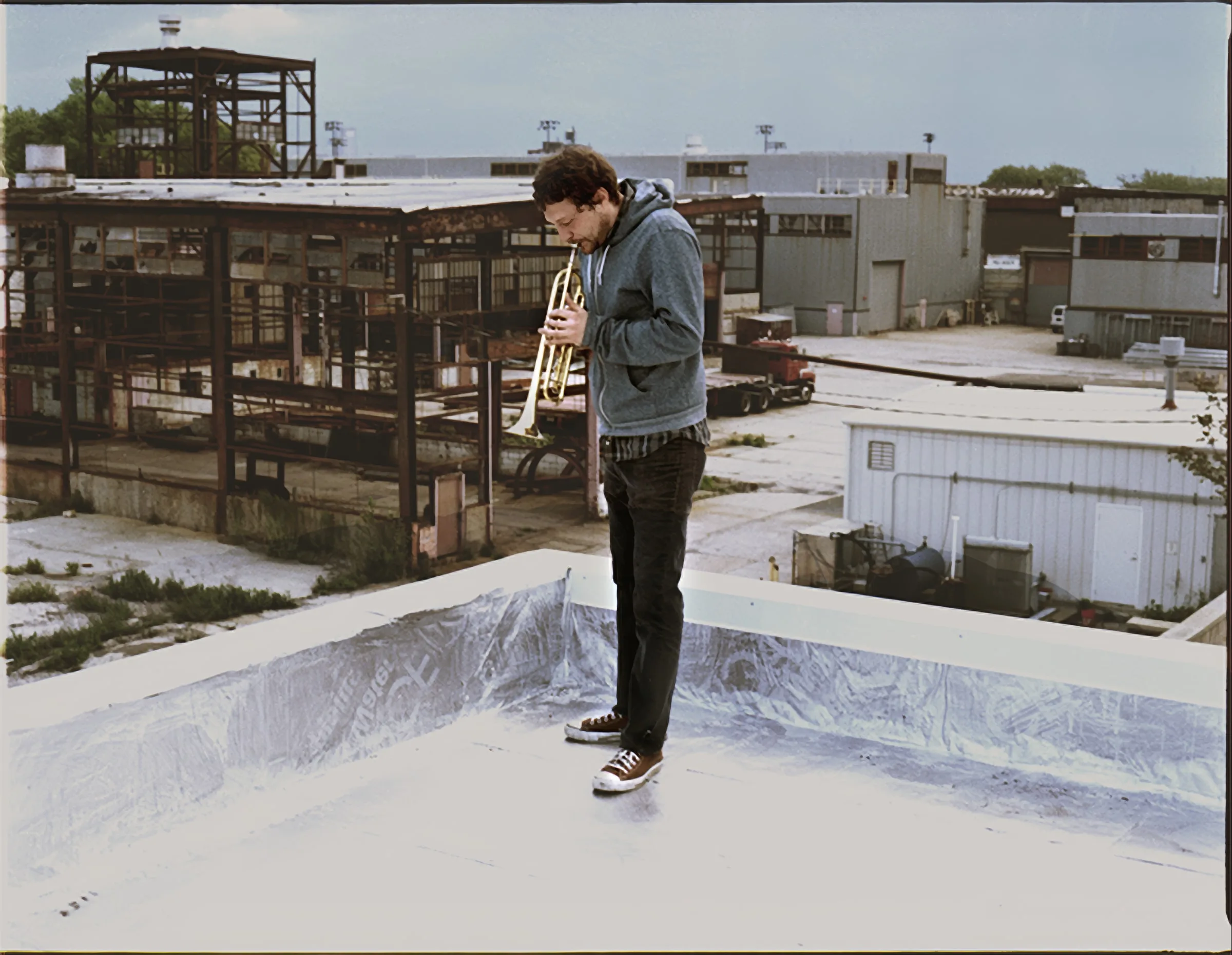A man in a gray jacket playing a trumpet on a white rooftop among old industrial buildings